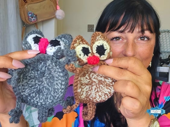 Ariana holds two crochet splattered cat coasters, one gray and one brown, toward the camera to compare the color options. Each coaster has large eyes, floppy ears, a bright pink nose, and curled paws. Original design by Ariana of CraftingHappiness.