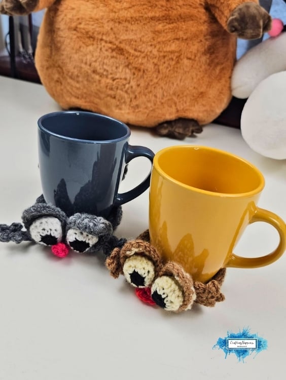 Gray and brown crochet splattered cat coasters wrapped around a dark blue mug and a mustard yellow mug, showing how they fit snugly around the base of each cup. The playful cats have oversized eyes, floppy ears, and bright pink noses. Original design by Ariana of CraftingHappiness.