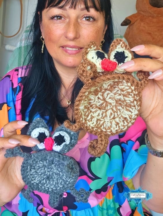 Ariana holds a gray and a brown crochet splattered cat coaster, showing both color variations side by side. The round coasters have oversized eyes, floppy ears, curly paws, and bright pink noses, making them look like flattened cartoon cats. Original design by Ariana of CraftingHappiness.