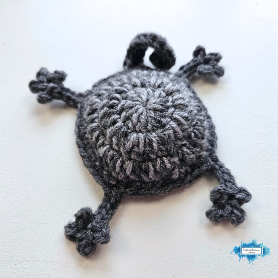 The gray crochet body of the Splattered Cat Coaster lies flat on a light surface, showing its round shape with four stretched paws and a curled tail before the face pieces are attached. Original design by Ariana of CraftingHappiness.
