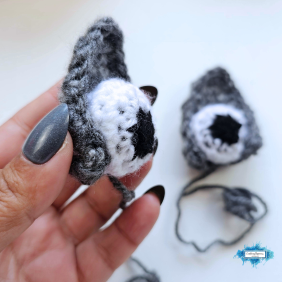 A hand wraps a gray crocheted ear around a white and black stuffed eyeball to create a cocoon effect. Original design by Ariana of CraftingHappiness.
