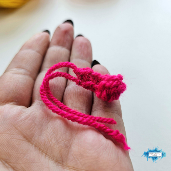 Two completed crochet eyes made with white yarn and black pupils are held between both hands. The gray coaster body and other crochet pieces are visible blurred in the distance, showing the parts before assembly. Original design by Ariana of CraftingHappiness.