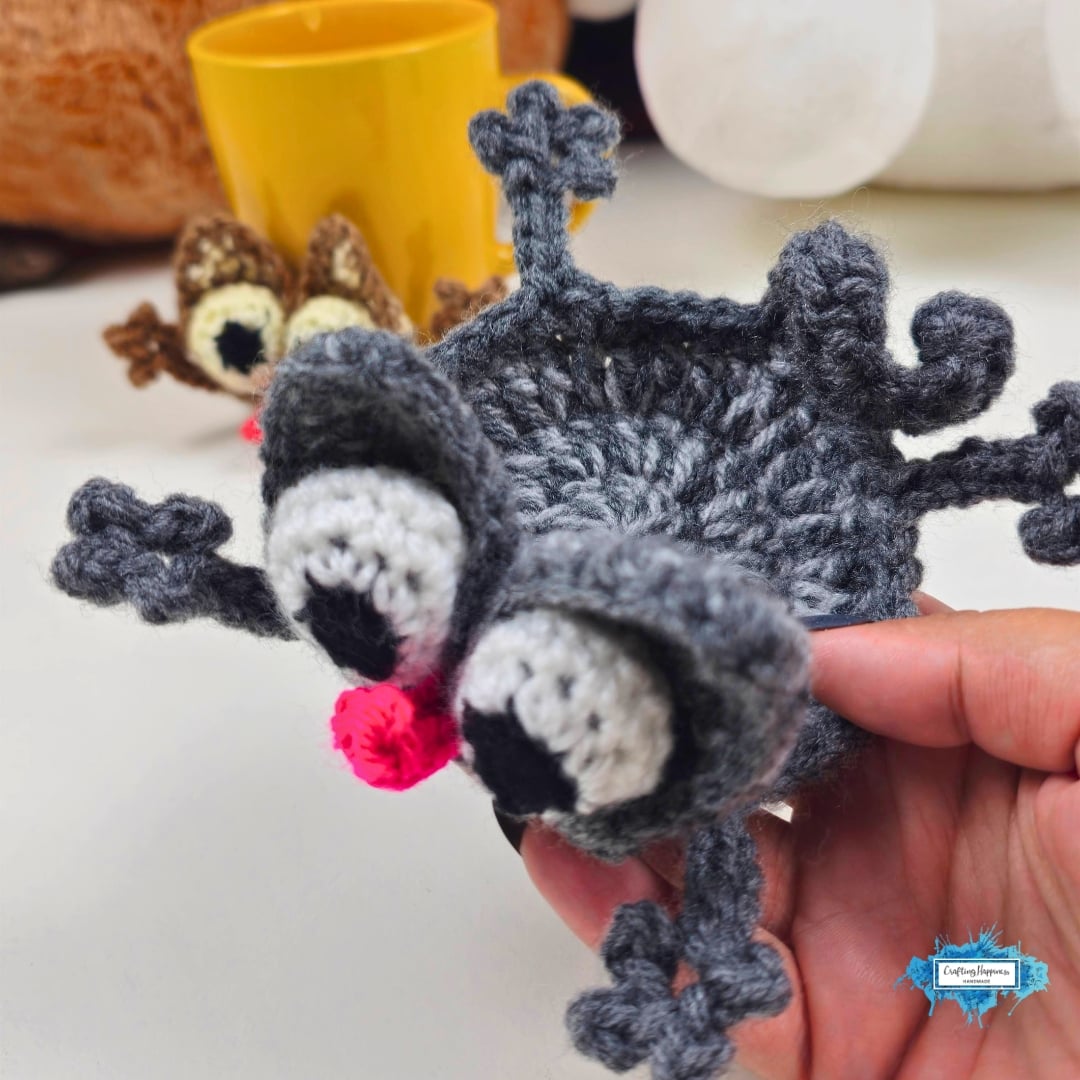 Close-up of a gray crochet splattered cat coaster held in one hand, highlighting its textured body, oversized eyes, pink nose, floppy ears, and stretched paws. A brown version and yellow mug appear softly blurred in the distance. Original design by Ariana of CraftingHappiness.