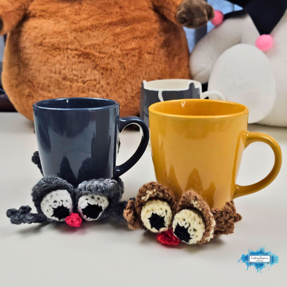 Gray and brown crochet splattered cat coasters wrapped around a dark blue mug and a mustard yellow mug on a tabletop. Both cats have large eyes, floppy ears, pink noses, and spread-out paws, showing how the coaster hugs the base of each mug. Original design by Ariana of CraftingHappiness.