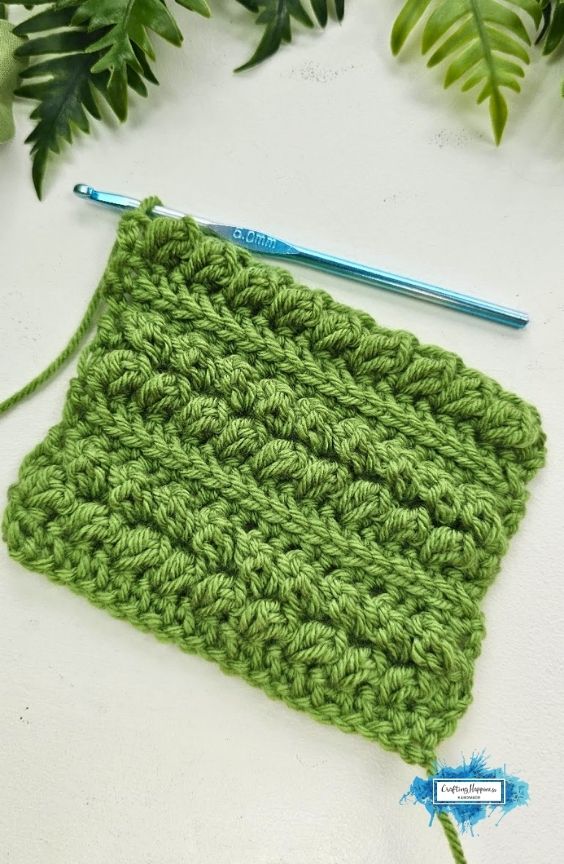 A green crochet swatch shows several completed rows of the Braided Leaves Stitch with raised leafy texture and a blue crochet hook resting at the top. The Braided Leaves Stitch was created by Ariana Wimsett, a known crocheter and author, and the crochet work shown here was also made by her.
