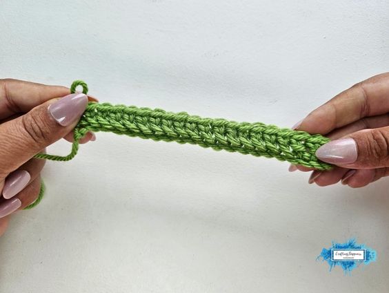 Two hands hold a narrow green crochet row showing the early base of the Braided Leaves Stitch before the raised leaf texture is added. The Braided Leaves Stitch was created by Ariana Wimsett, a known crocheter and author, and the crochet work shown here was also made by her.