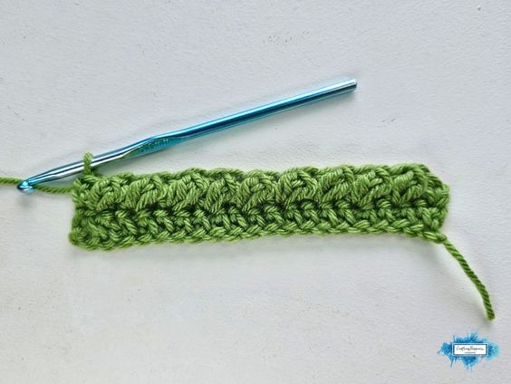 A narrow green crochet swatch shows Row 3 of the Braided Leaves Stitch with raised textured sections forming along the row and a blue crochet hook resting above it. The Braided Leaves Stitch was created by Ariana Wimsett, a known crocheter and author, and the crochet work shown here was also made by her.