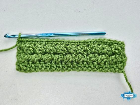 A green crochet swatch shows Row 4 of the Braided Leaves Stitch with a completed textured leaf row and a blue crochet hook resting across the top. The Braided Leaves Stitch was created by Ariana Wimsett, a known crocheter and author, and the crochet work shown here was also made by her.