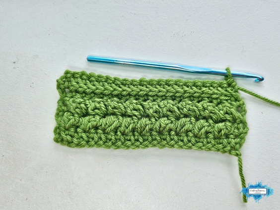 A green crochet swatch shows Row 5 of the Braided Leaves Stitch, with raised leaf-like texture forming across the fabric and a blue crochet hook resting along the top edge. The Braided Leaves Stitch was created by Ariana Wimsett, a known crocheter and author, and the crochet work shown here was also made by her.